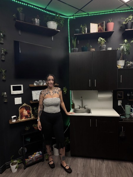 Davi standing in her Pretty Ugly studio in Charlotte, surrounded by plants on dark shelves